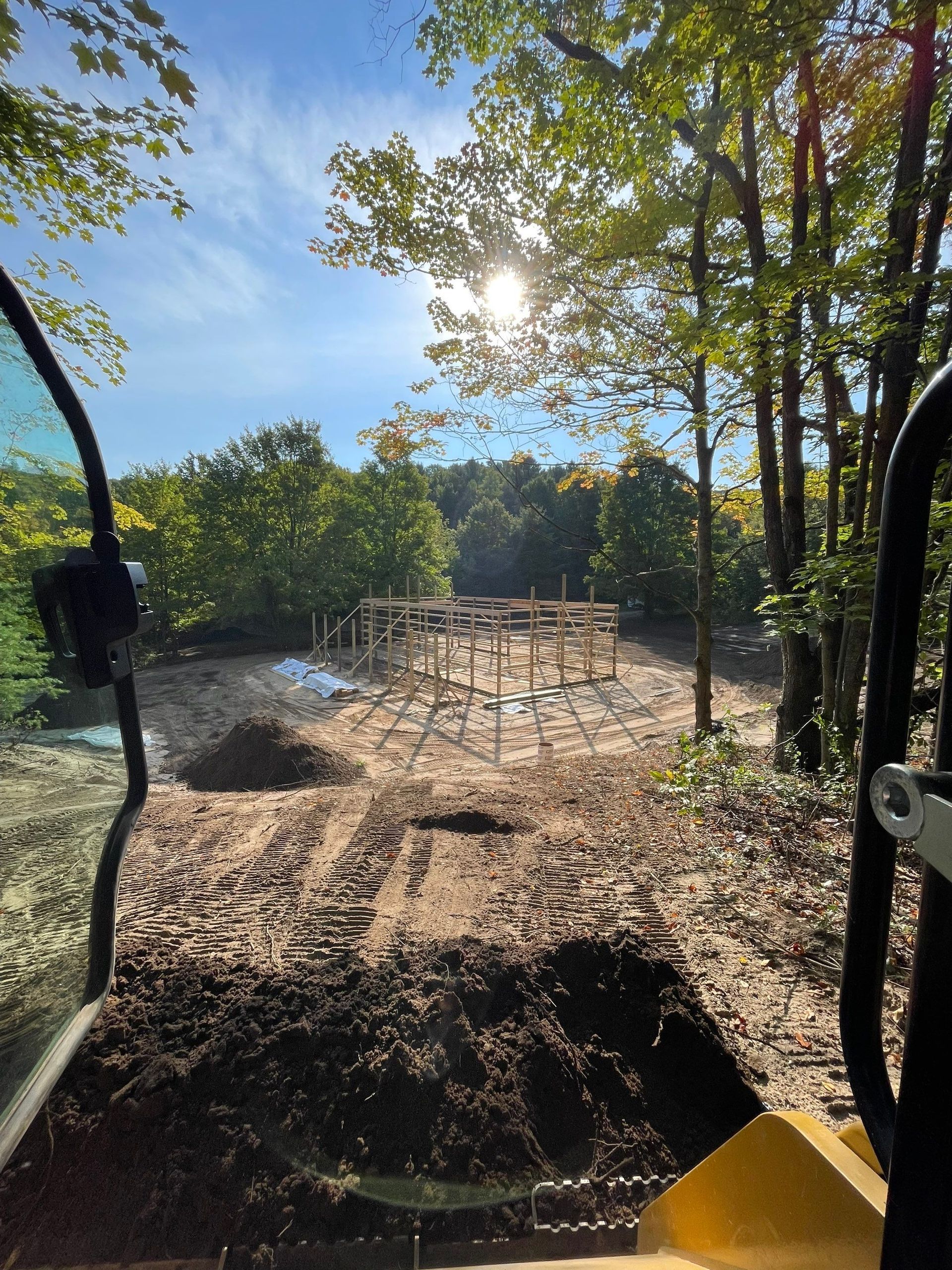 Construction site in a wooded area with framework and equipment. Bright sunlight through trees, blue sky.