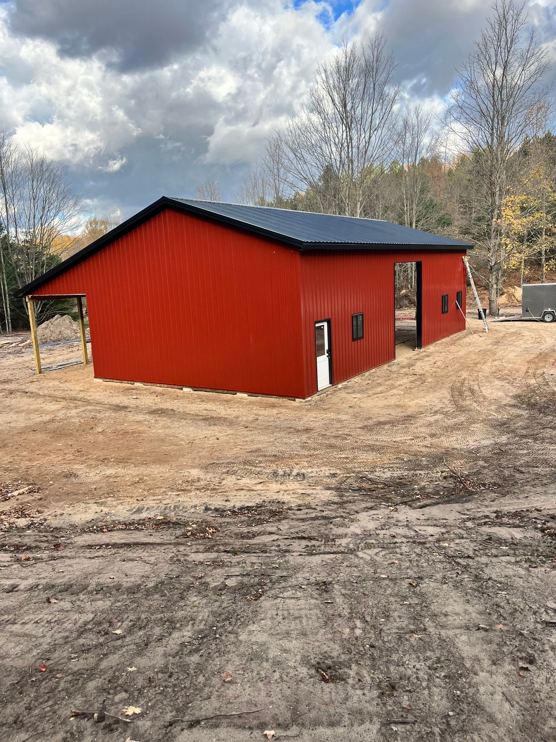 Red metal barn with a black roof and open doorways on a dirt lot under a cloudy sky.
