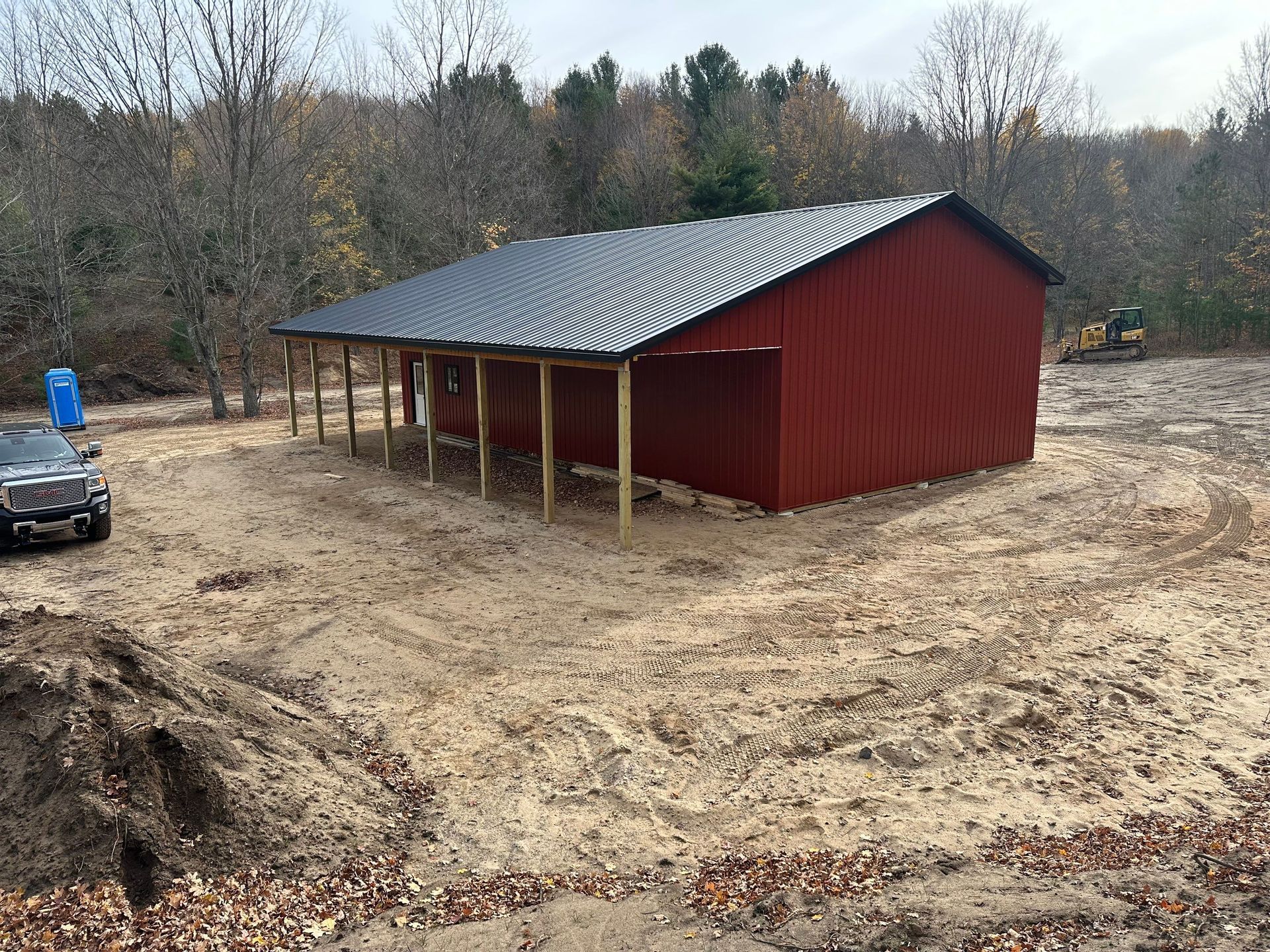 Red barn with black roof, supported by wooden posts, on a sandy lot surrounded by trees.