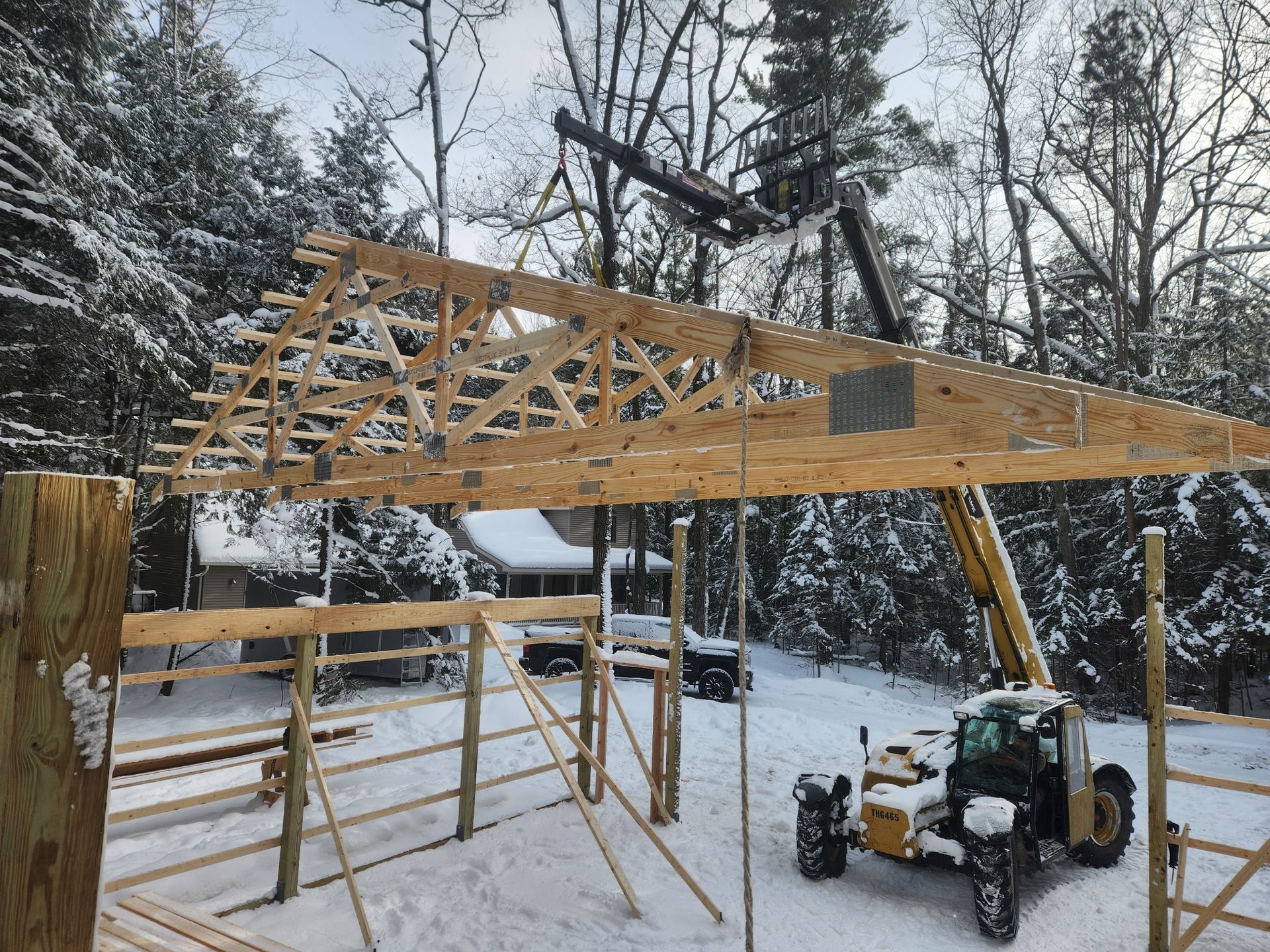 A large wooden roof truss being lifted into place by a yellow telehandler on a snowy construction site.
