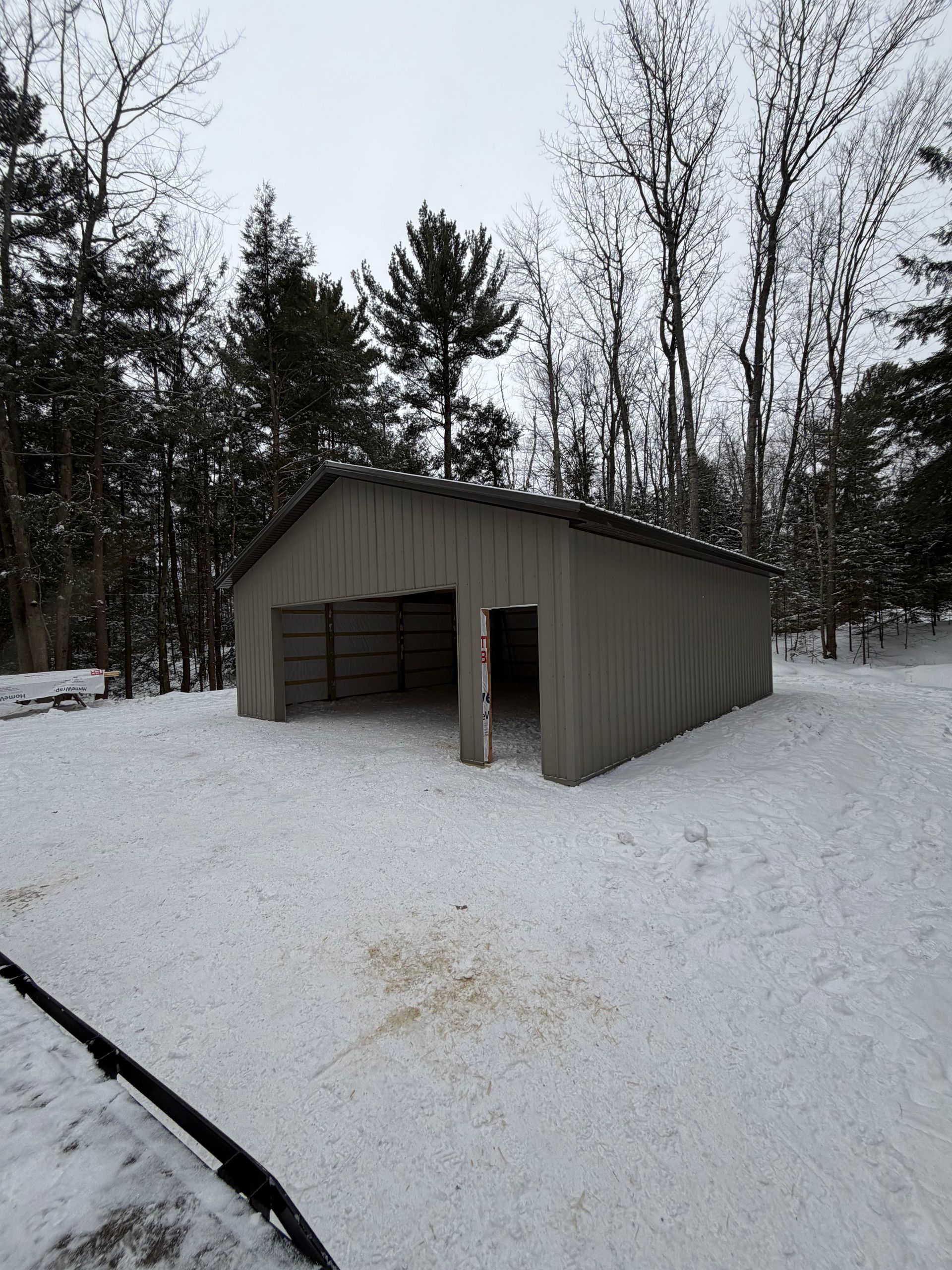 A tan open-sided shed in a snowy, wooded area.