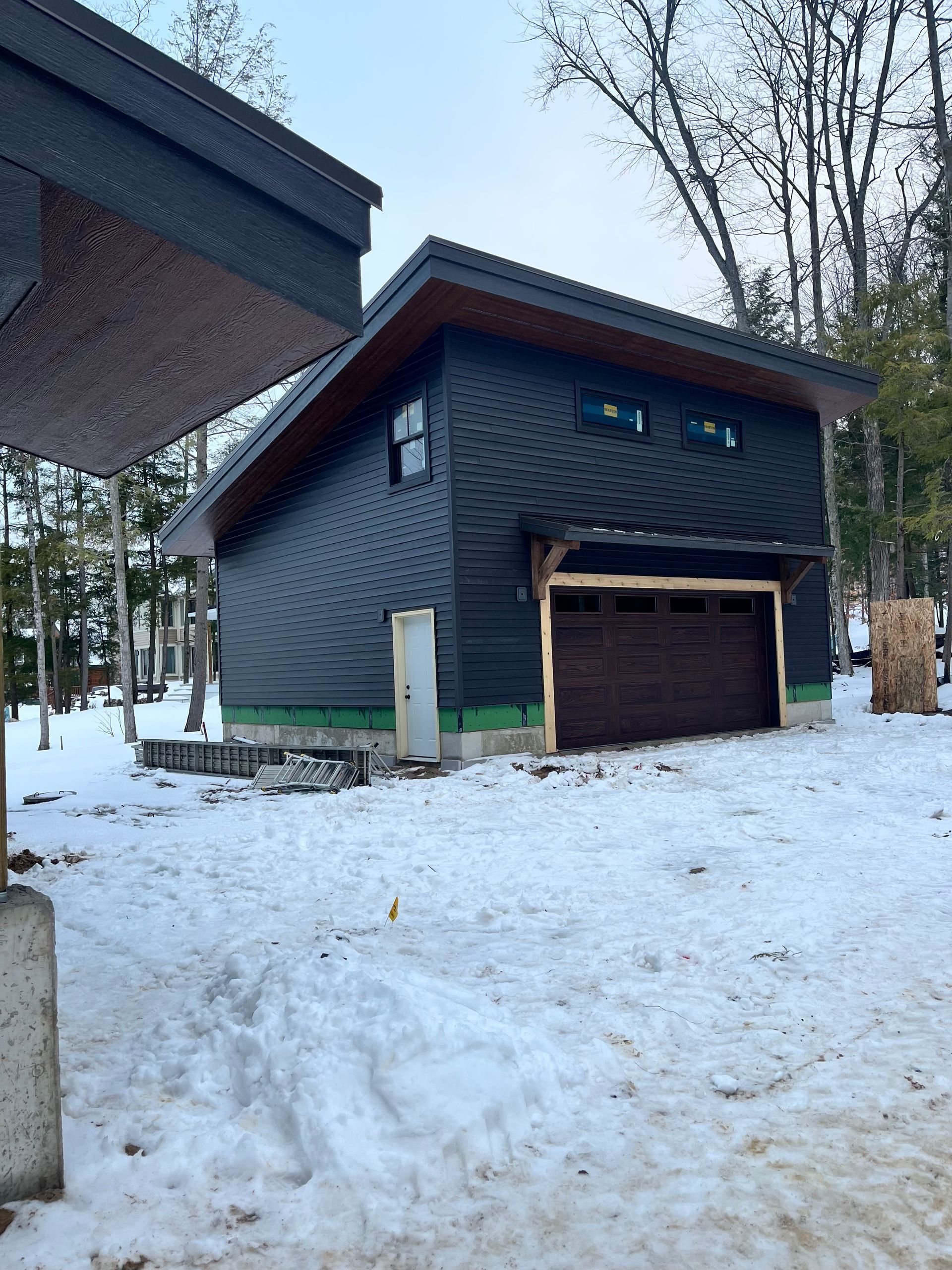 Modern, dark-sided building with a brown garage door, set in a snowy, wooded area.