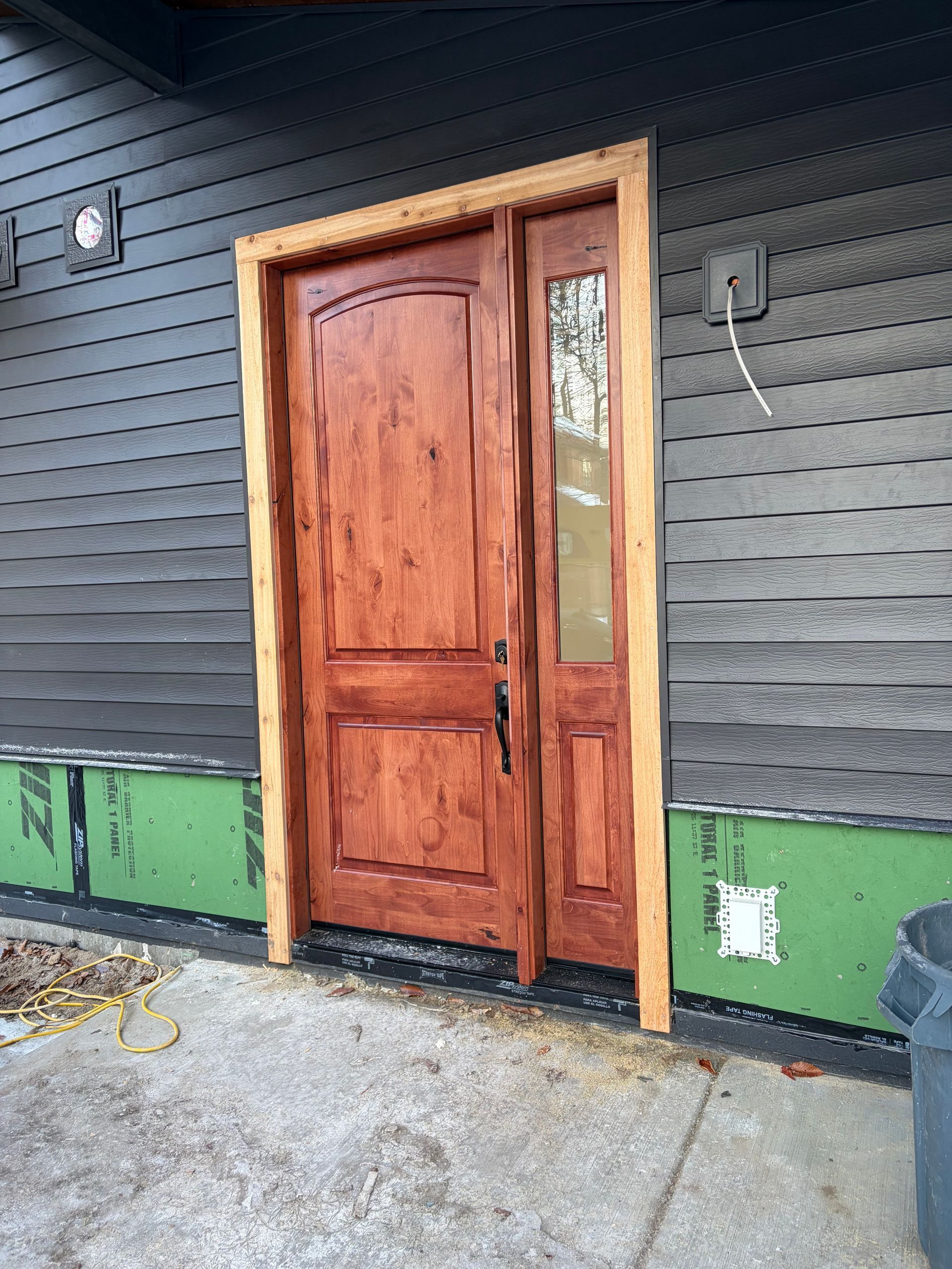 Brown wooden front door with sidelight, tan trim, dark gray siding, concrete slab.