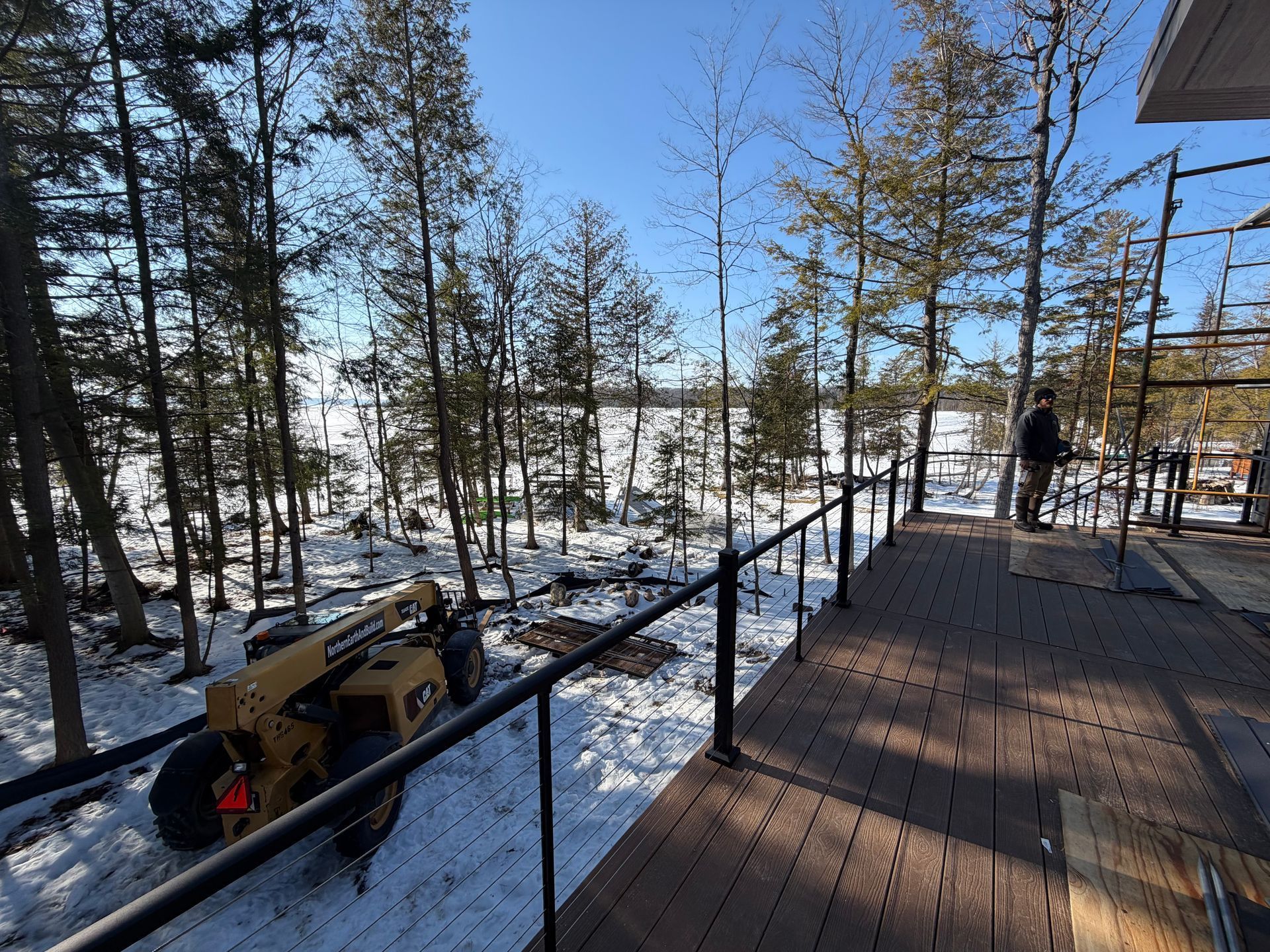Yellow construction vehicle on snowy landscape next to wooden deck with black railing, trees, and person.
