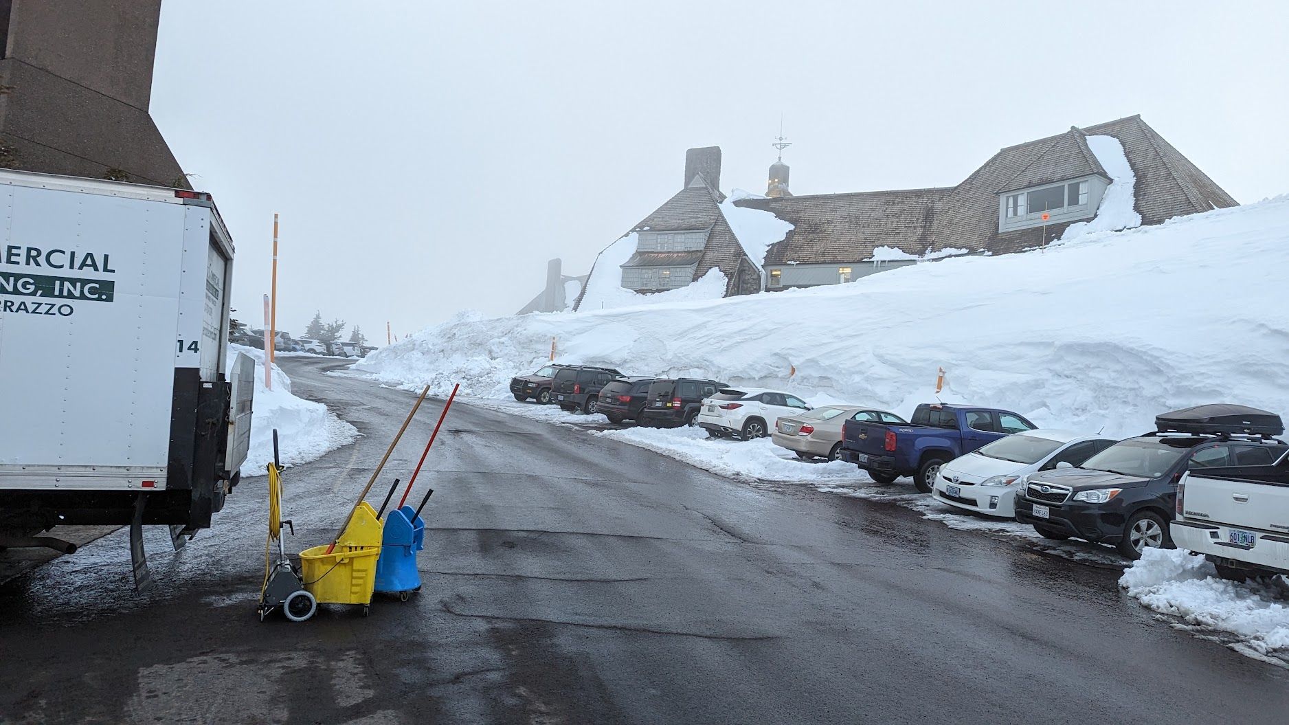 Snowy parking lot beside buildings, with parked cars, a truck, and shovels on a wet road.