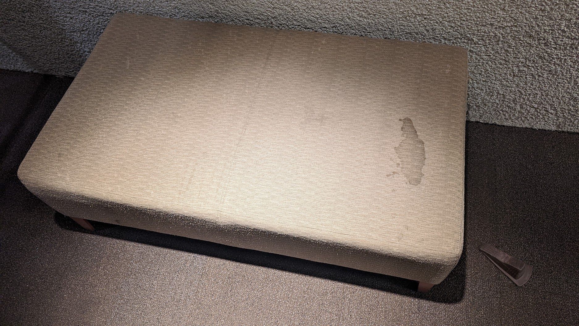Beige rectangular ottoman on carpeted floor, with a small tear on the top.