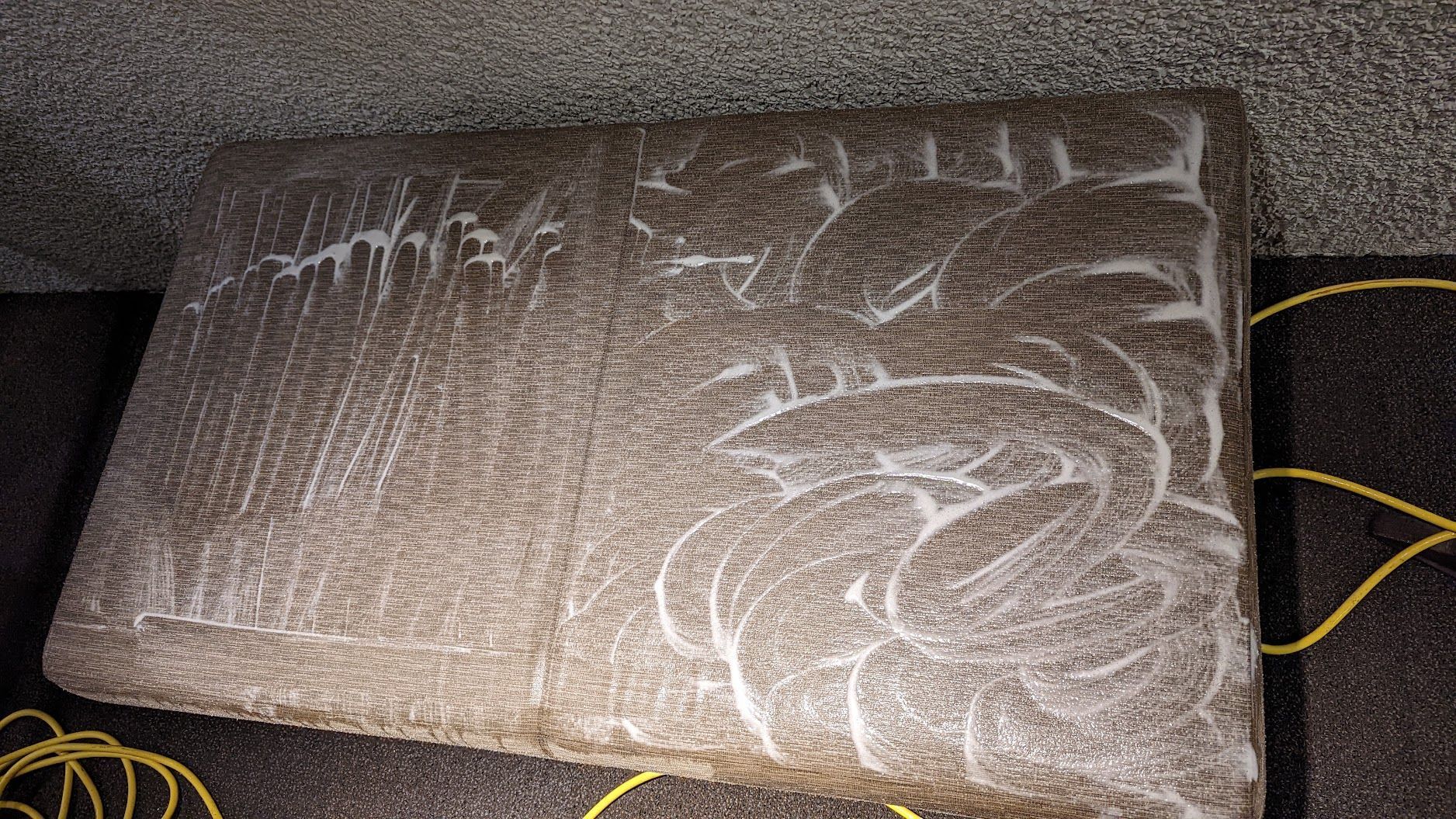 Brown cushion or mattress with white leaf-like patterns on a carpet, with yellow cords nearby.