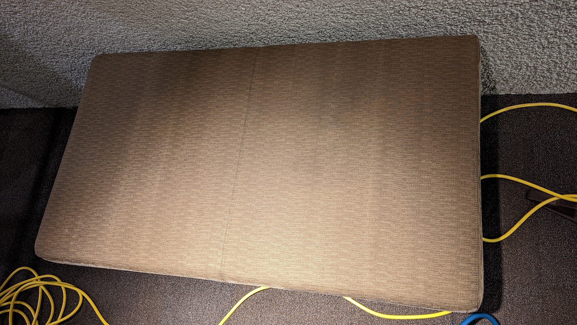 Tan rectangular cushion or pad on a carpeted floor with yellow cables nearby
