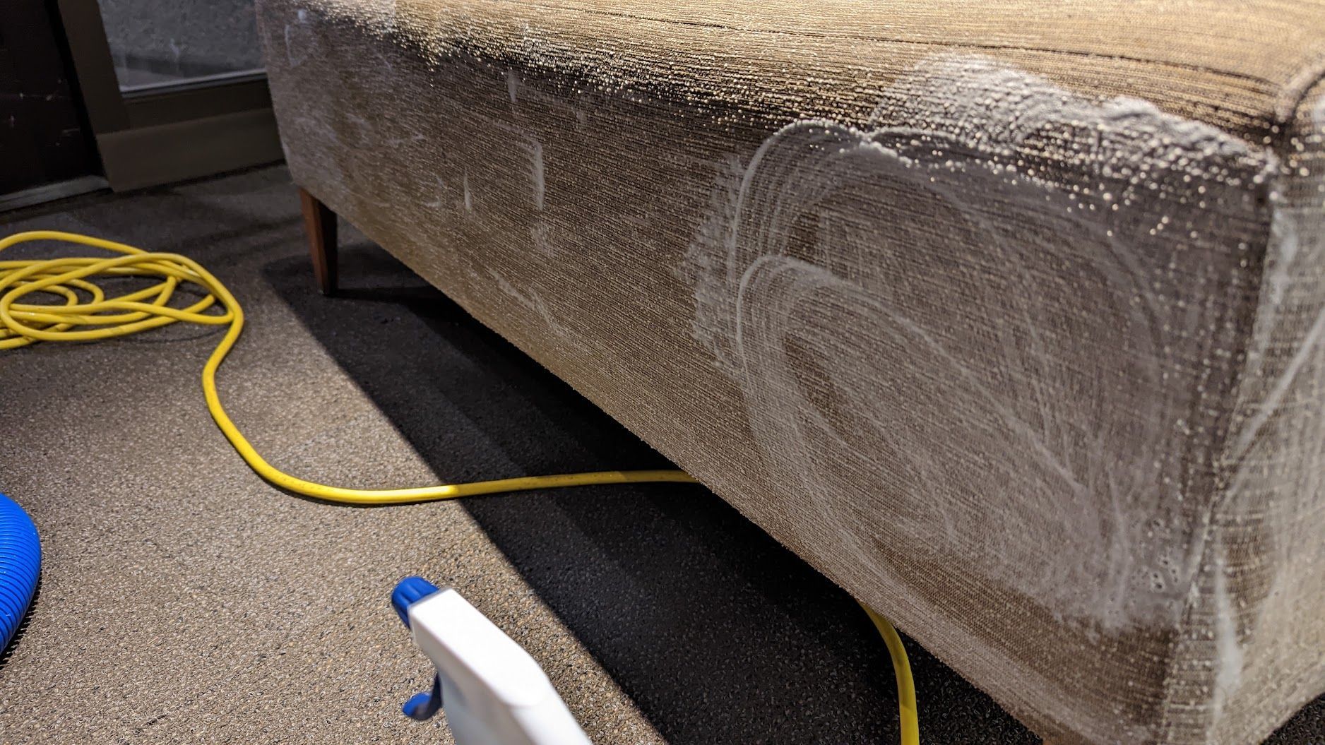 Couch wrapped in clear plastic with a yellow cord and spray bottle on carpeted floor