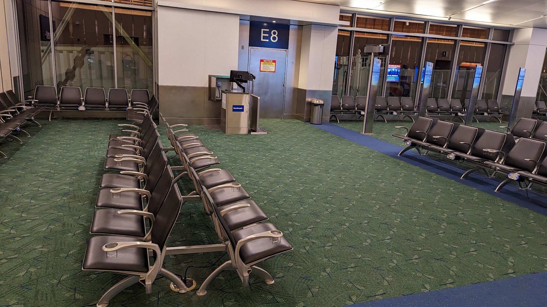 Empty airport gate seating area with rows of black chairs on green carpet near a blue E8 sign.