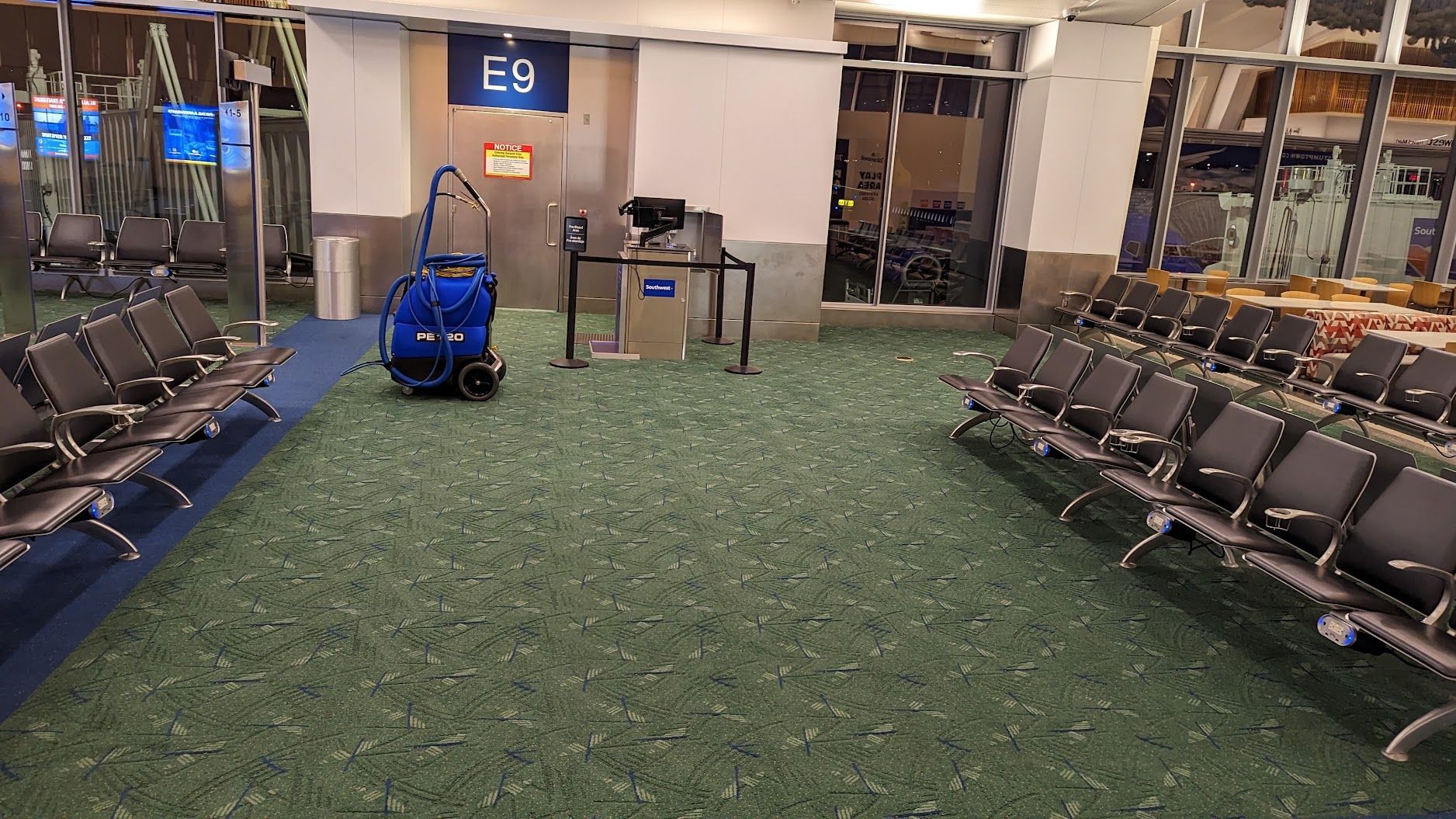 Empty airport waiting area with rows of seats and a blue cart on a green carpet near gate E9