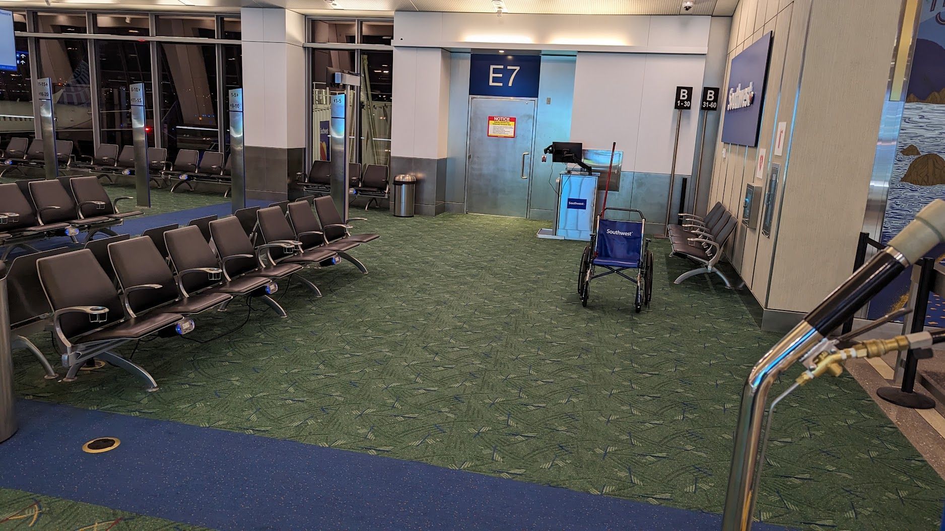 Empty airport gate area with rows of chairs, green carpet, blue wall, and luggage carts near gate E7