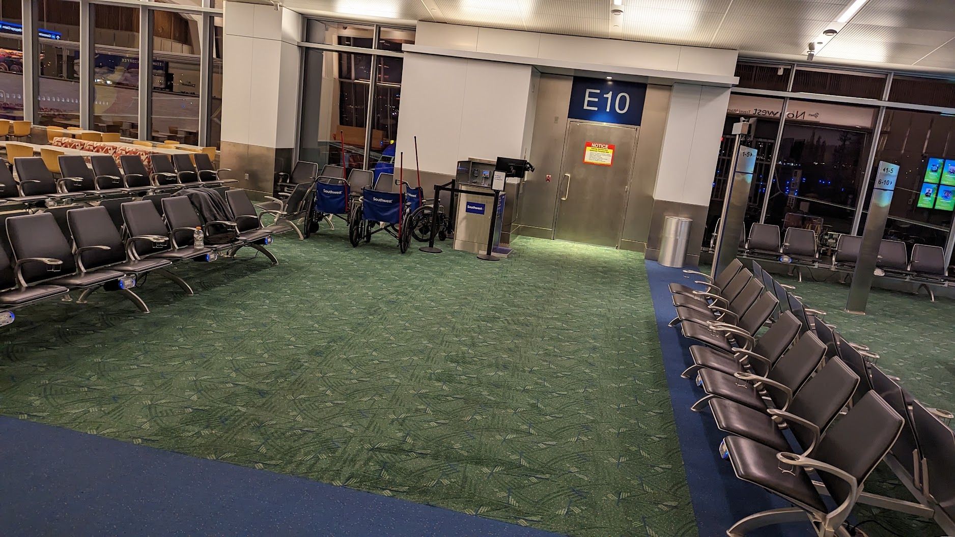 Empty airport gate seating area with rows of black chairs and green carpeted walkway.