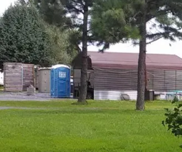 Blue portable toilet next to trees and wooden structures on a grassy area.