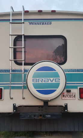 Rear view of a Winnebago Brave RV with a spare tire cover, ladder, and a license plate.