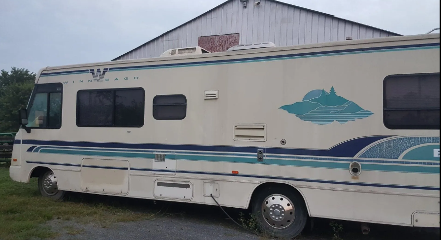 Cream-colored RV with blue and teal stripes and mountain graphic parked near a white building with a gray sky.