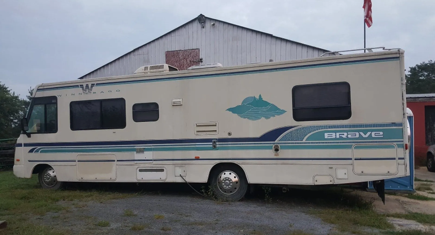 Cream-colored RV with blue accents parked in front of a white building with an American flag.