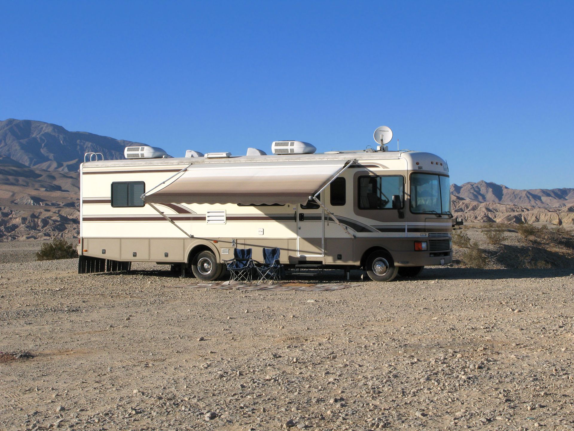 RV parked on gravel, awning extended, two chairs nearby. Mountains in the background, blue sky.