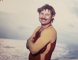 A man in a wetsuit is standing on the beach with his arms crossed.