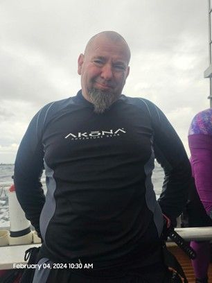 A man with a beard is wearing a black shirt with the word akena on it.