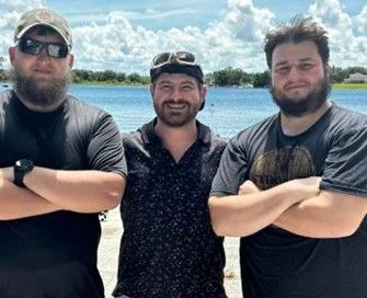 Three men are standing next to each other on a beach with their arms crossed.