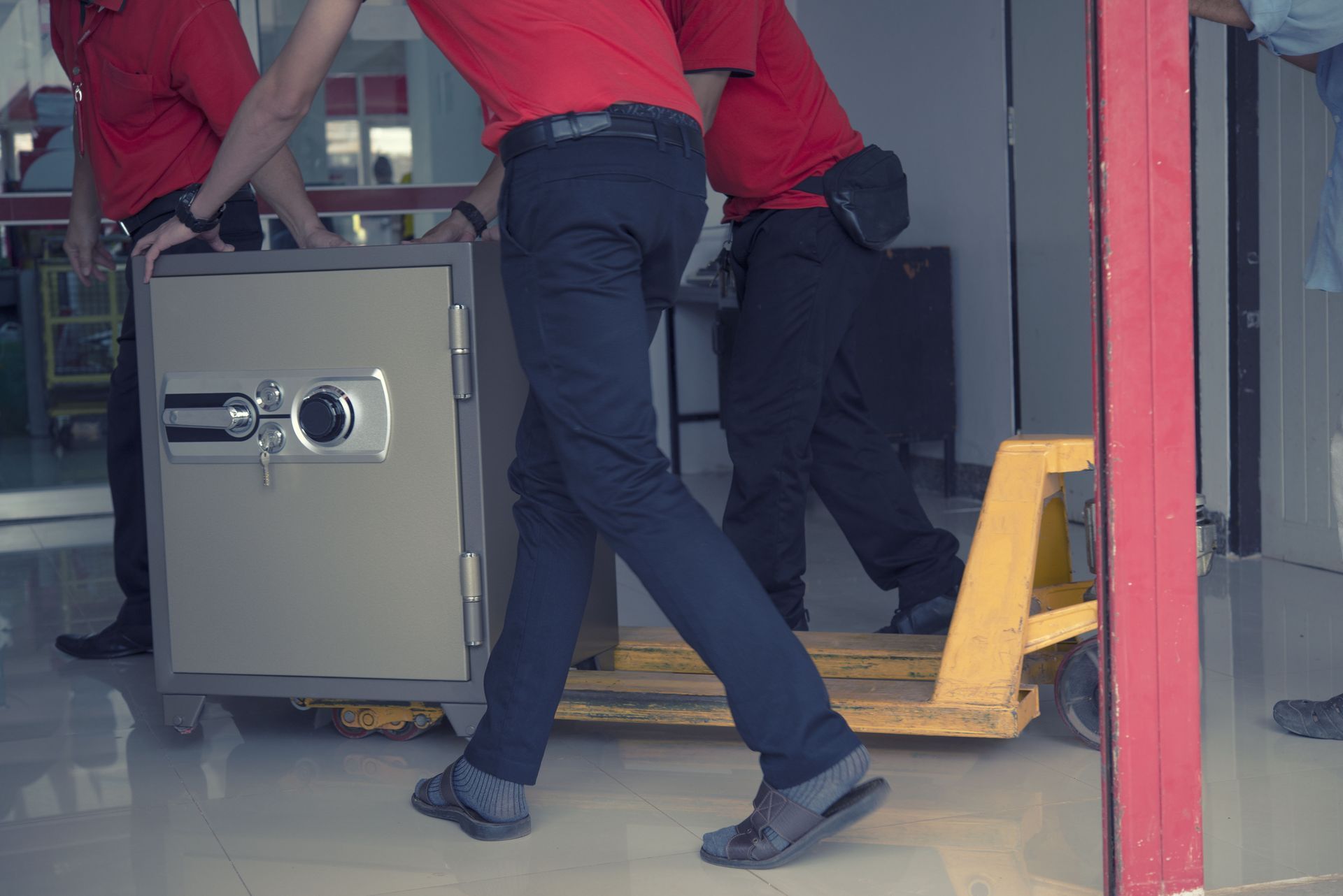Two workers in red shirts maneuver a large safe on a yellow pallet jack in a warehouse doorway