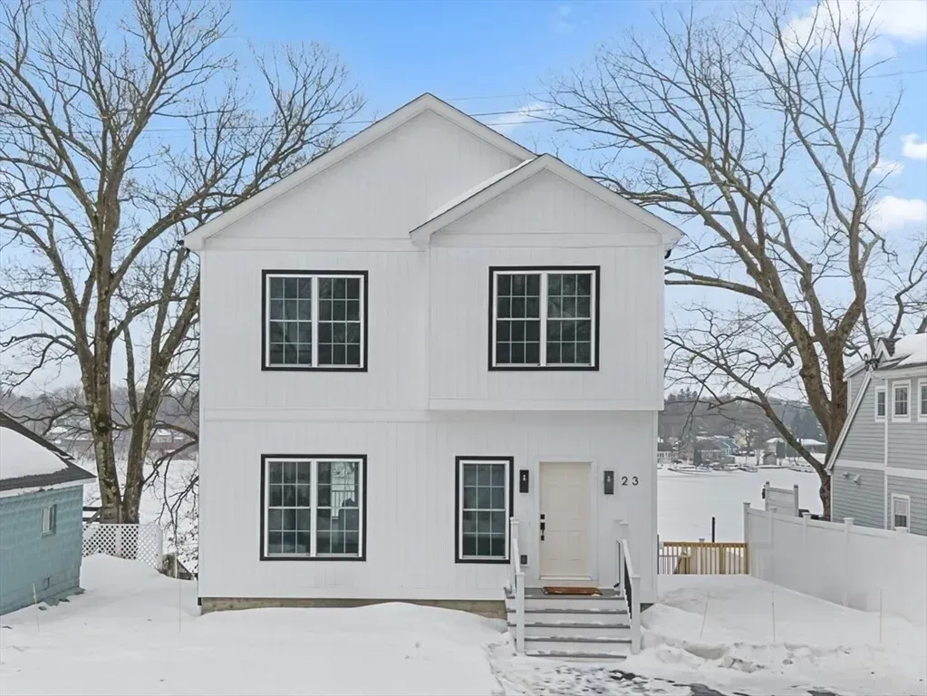 A two-story white house with dark window frames, surrounded by snow in a yard with bare trees in the background.