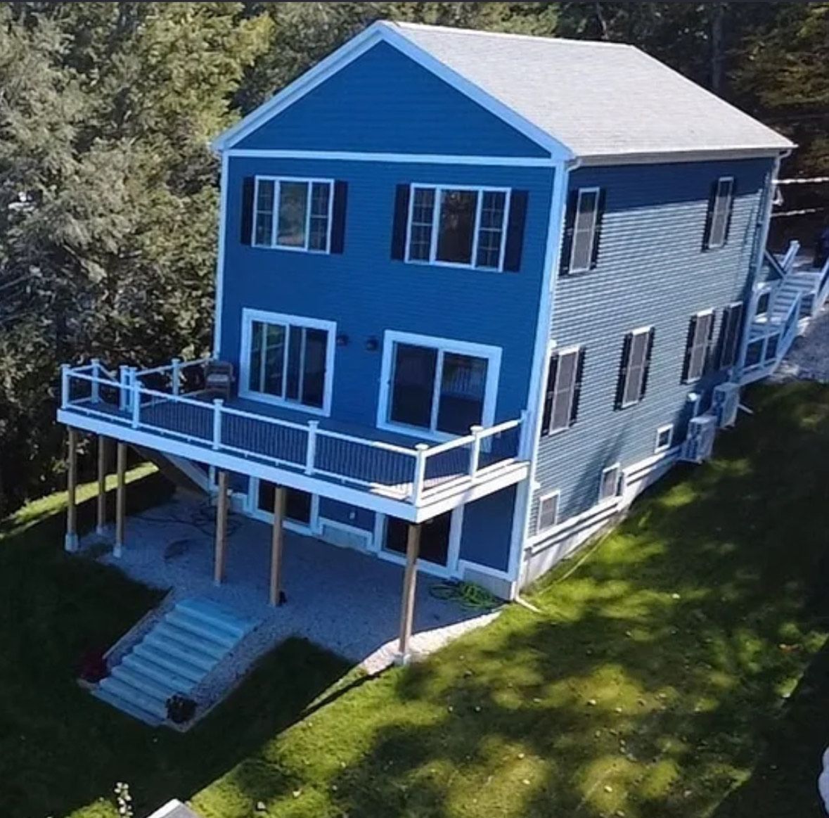 A multi-story blue house with white trim, a raised deck, and a stone patio, surrounded by green grass and trees.