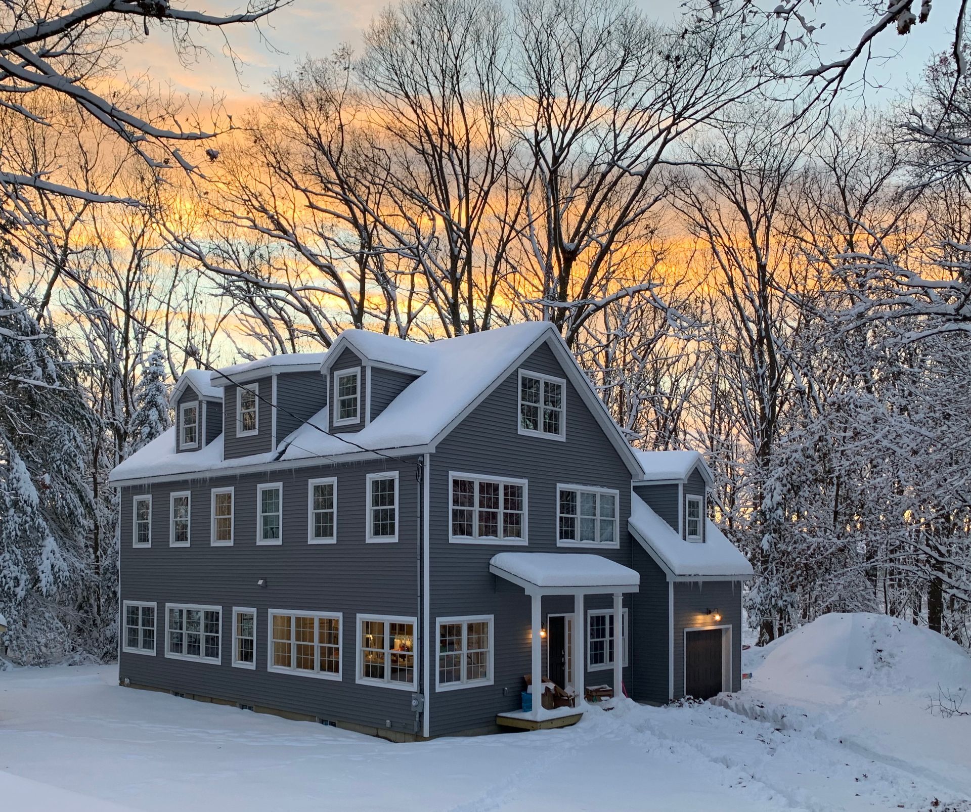 Gray house in snowy landscape at sunset. Windows lit, snow-covered trees and ground, orange and blue sky.