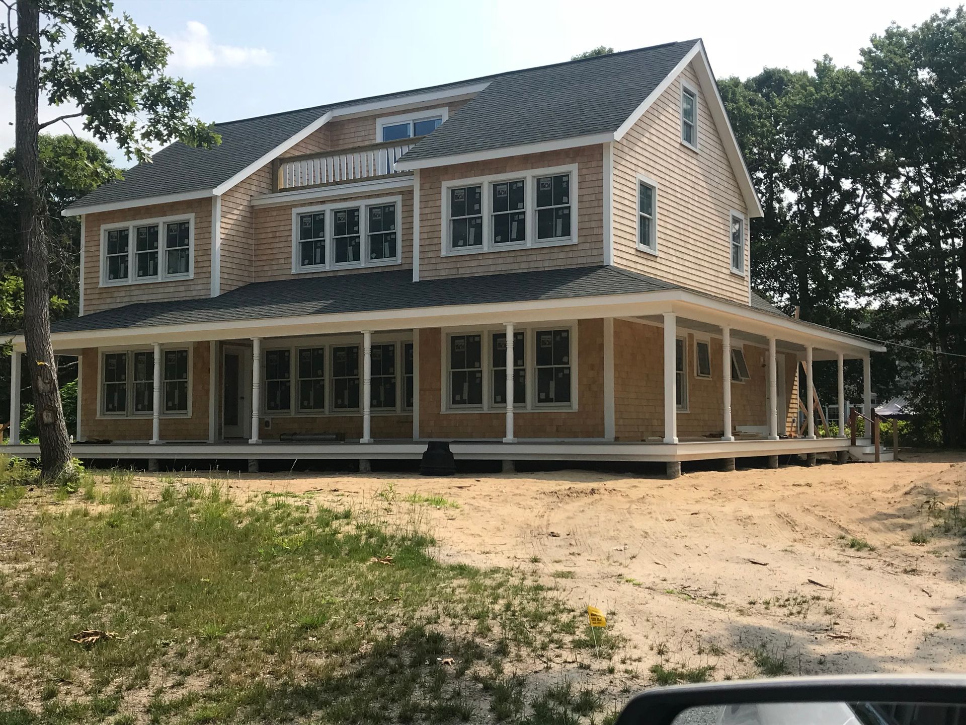 Two-story tan house with a wrap-around porch and dark roof, set in a sandy yard with some trees.