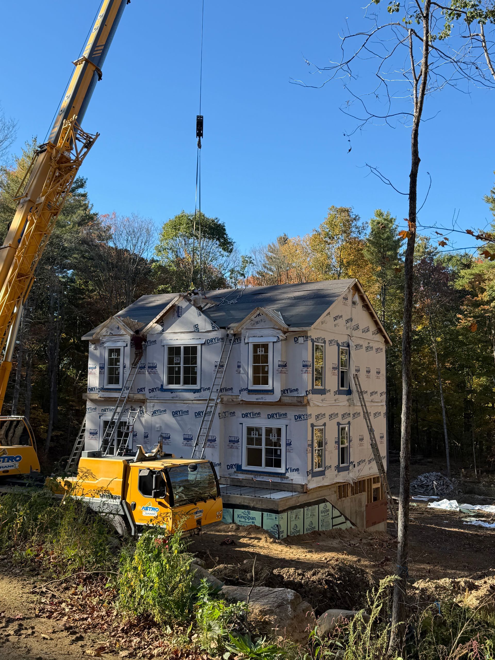 Crane lifting debris beside a damaged white house with a yellow truck in a wooded area under a clear blue sky