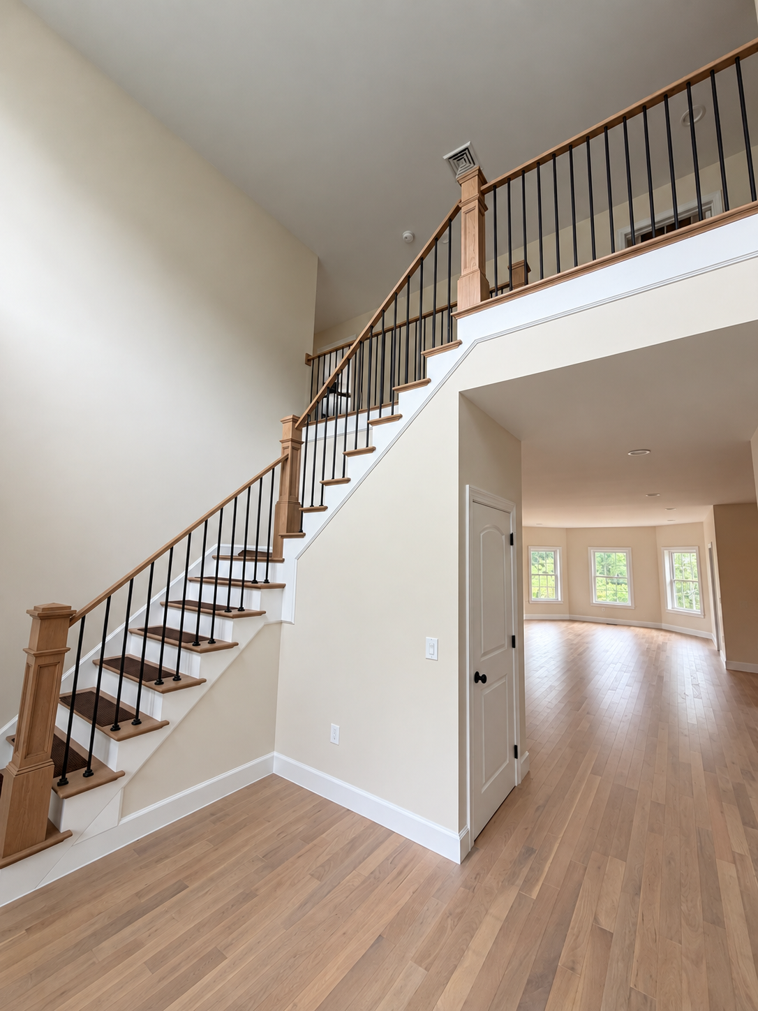 Interior staircase with wooden railings in a bright, empty home, opening to a living area with hardwood floors