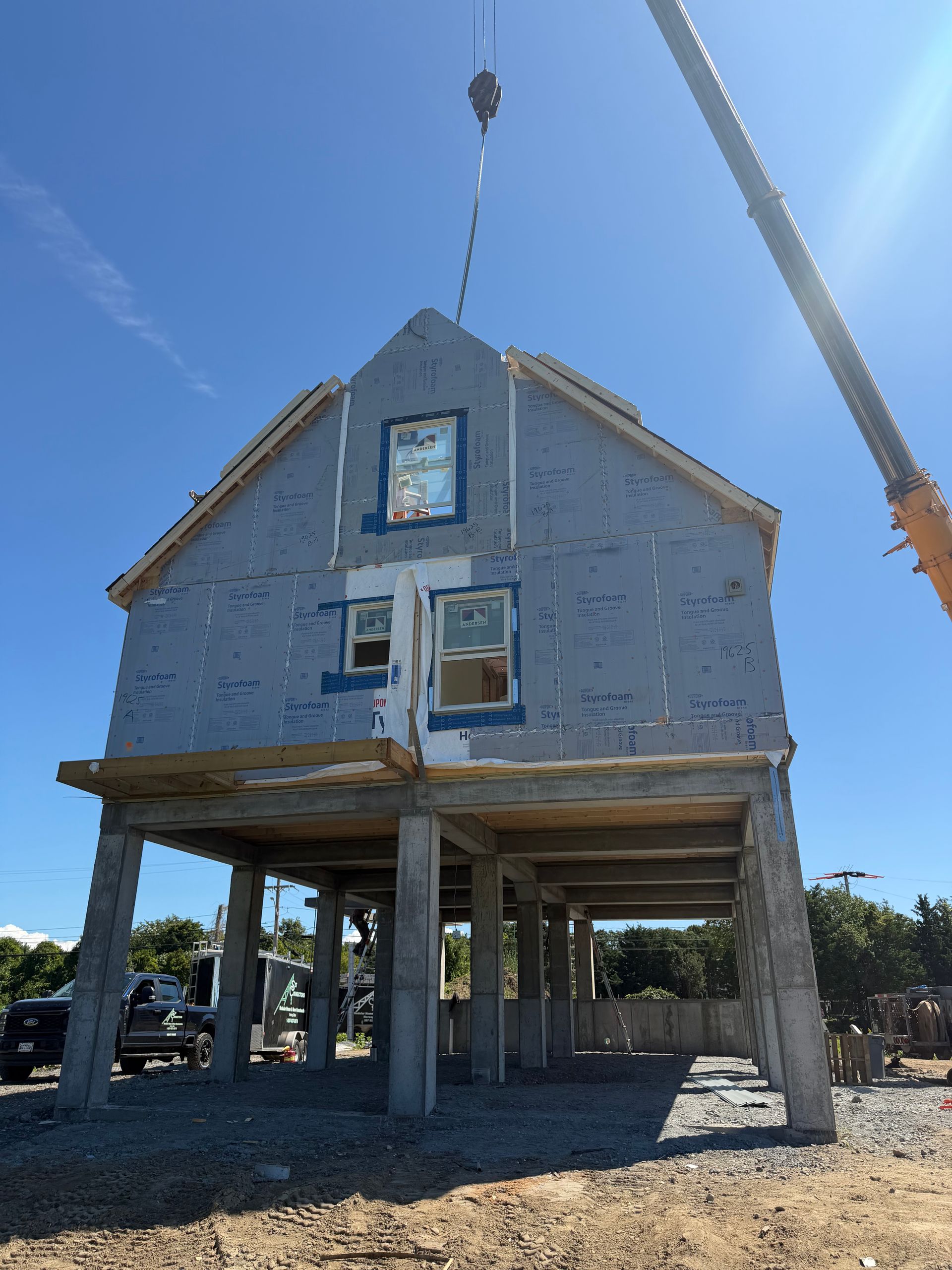 Stilted house under construction with exposed framing and a crane lifting a beam under a blue sky