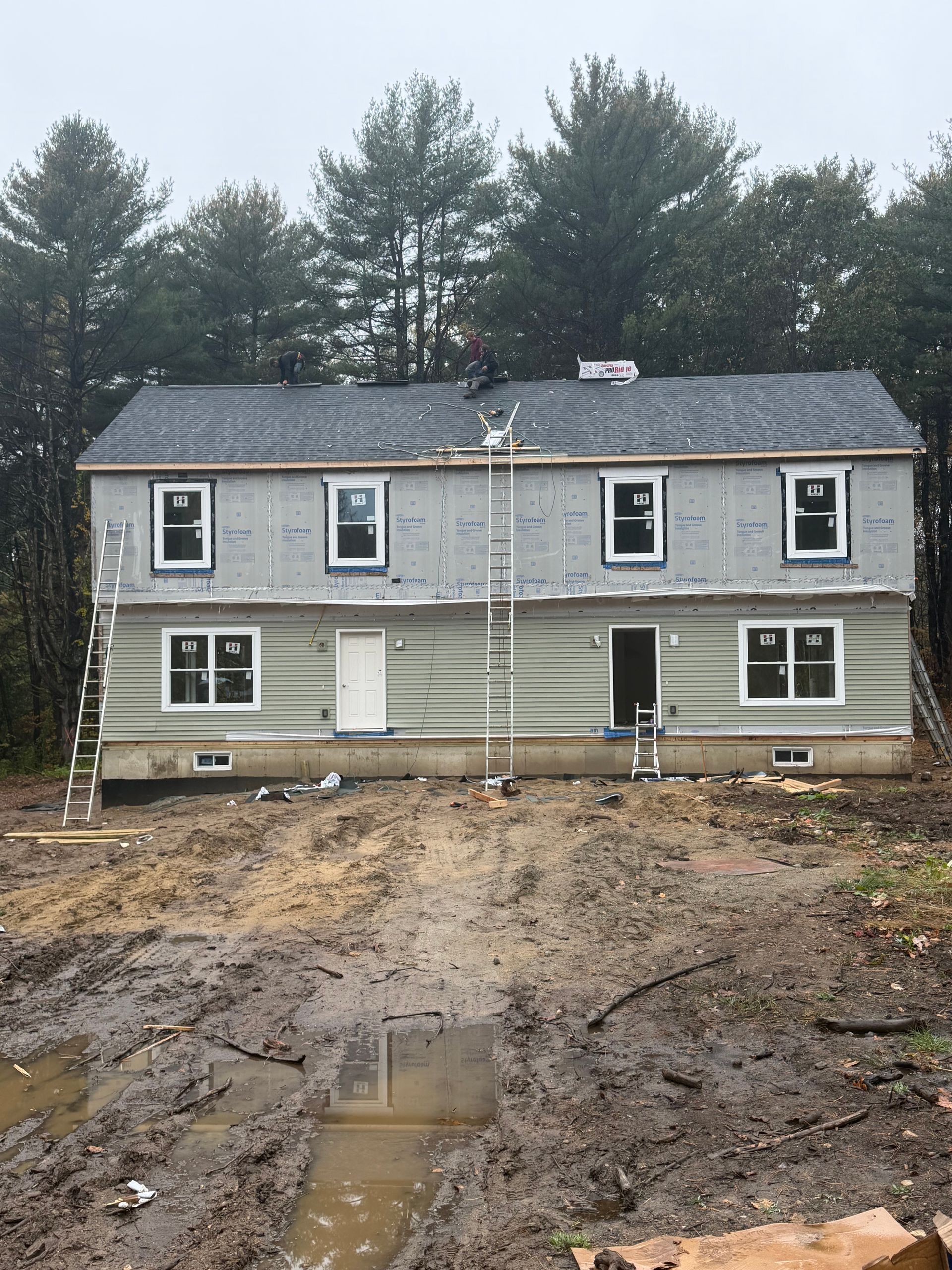 Two-story house with unfinished lower siding and muddy construction site in front, trees behind