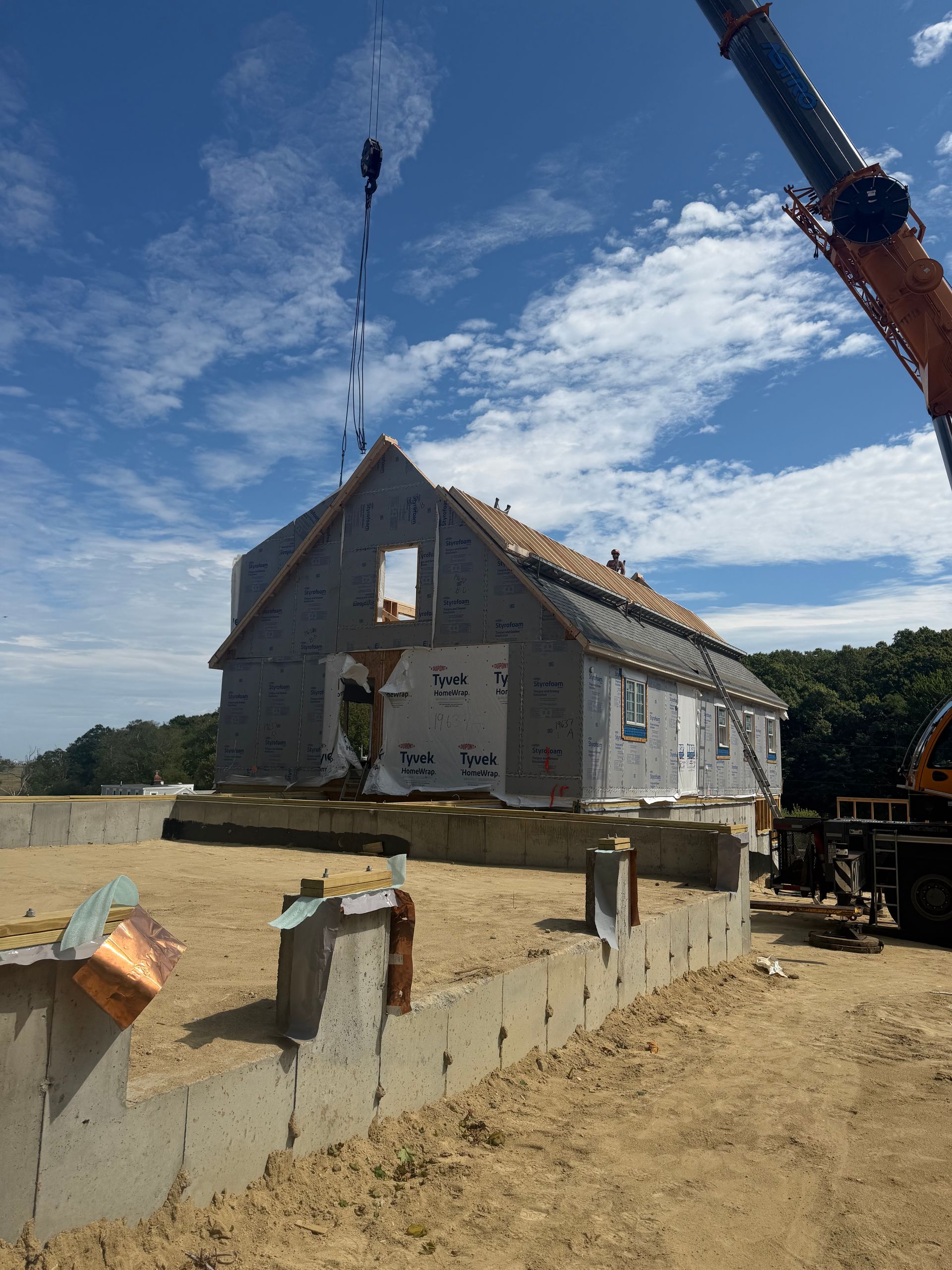 House being lifted by a crane, with a crane hook above the roof on a dirt lot under a blue sky.