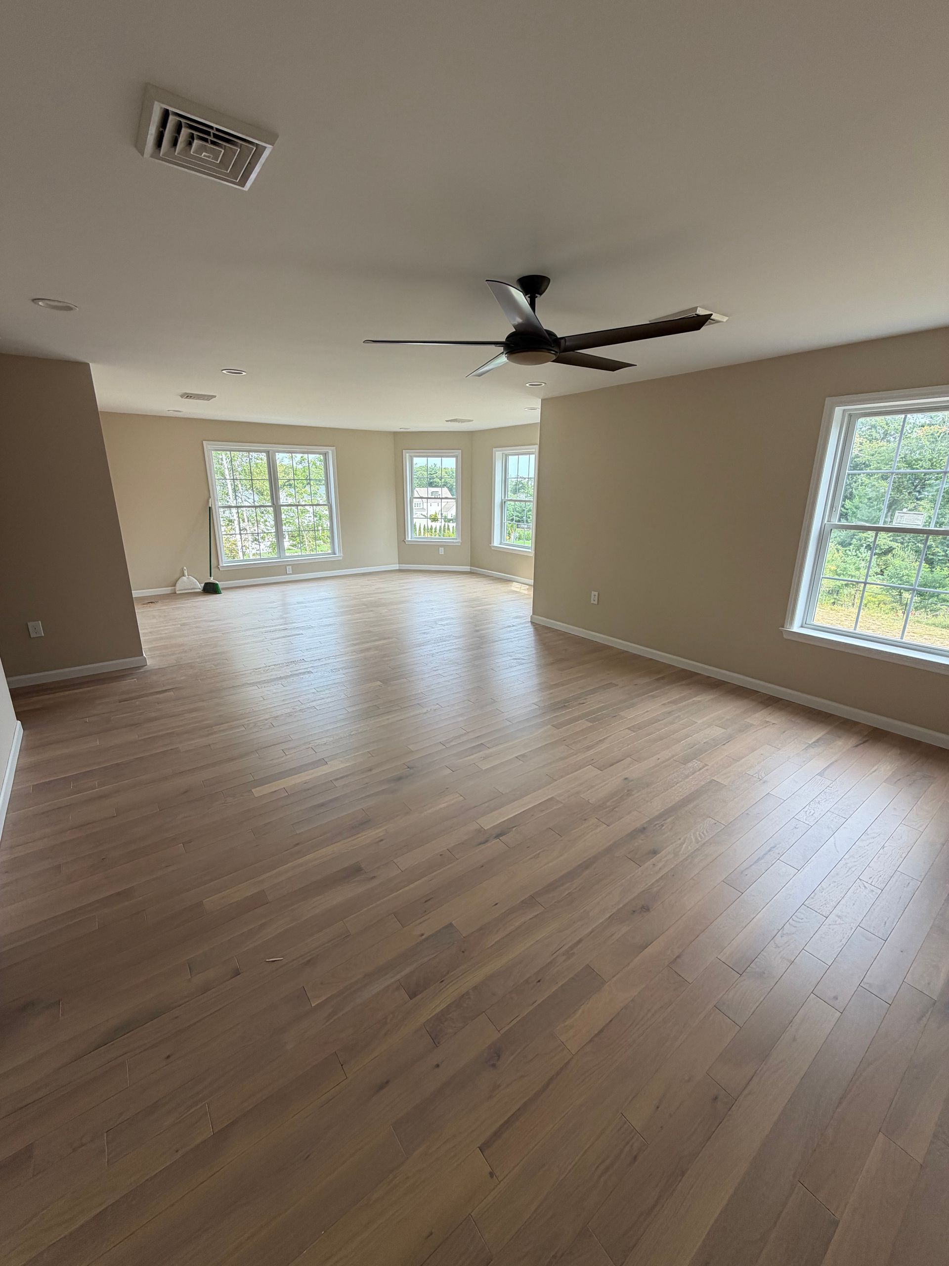 Empty beige living room with light wood floors, ceiling fan, and large windows.
