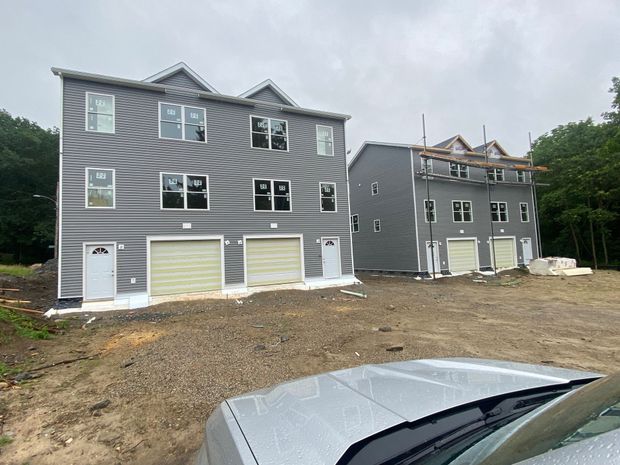 Two gray, multi-story townhouse buildings under construction with garages, surrounded by dirt and trees.