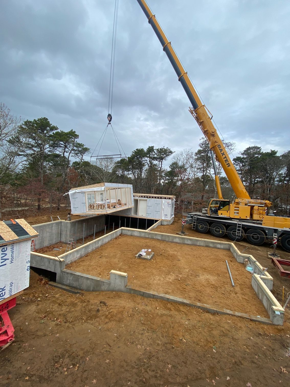 A crane lifting a modular home section above a concrete foundation on a construction site. Cloudy sky.