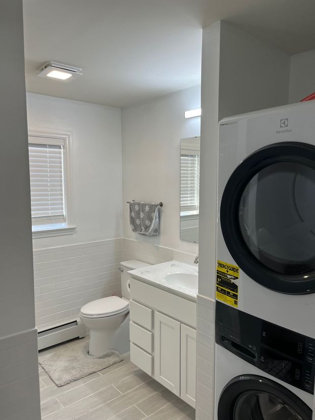 A white bathroom featuring a stacked washer and dryer, a vanity with a sink, a toilet, and a window with blinds.
