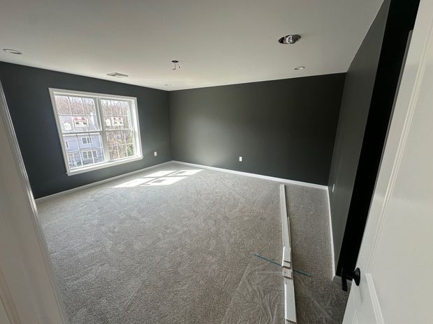 An empty room with grey walls, grey carpeting, a window on the left, and recessed ceiling lighting.