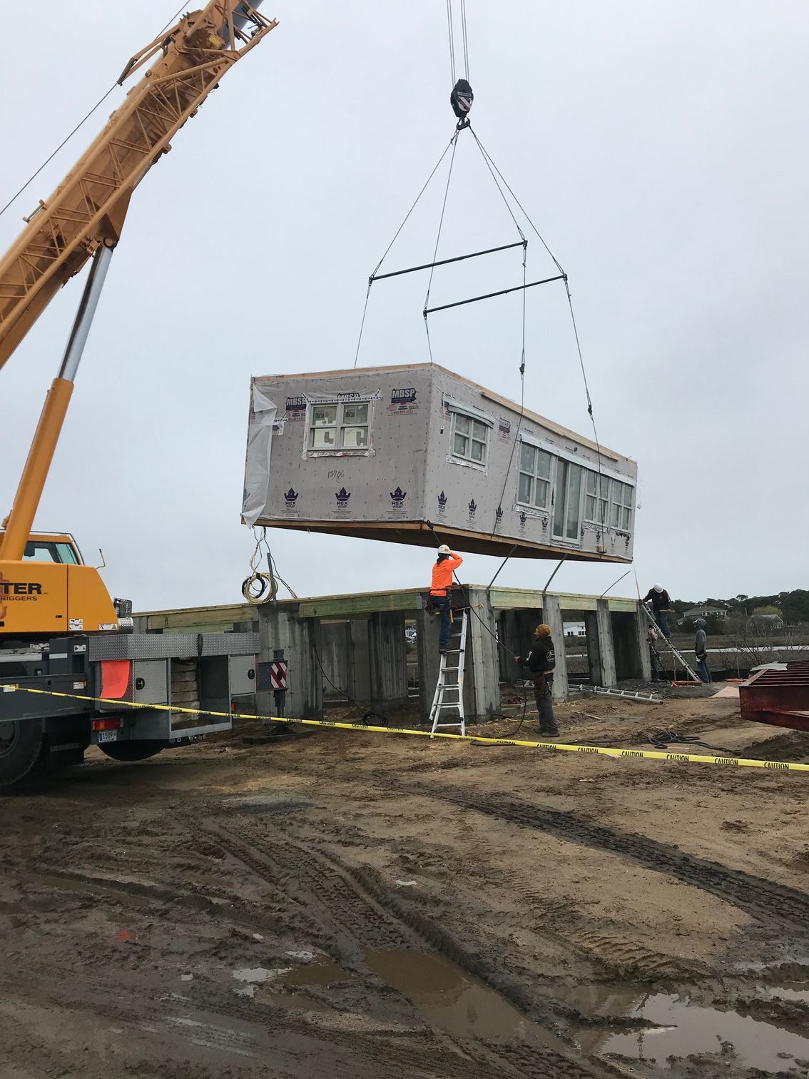 A crane lifting a modular building section into place at a construction site; two workers nearby.