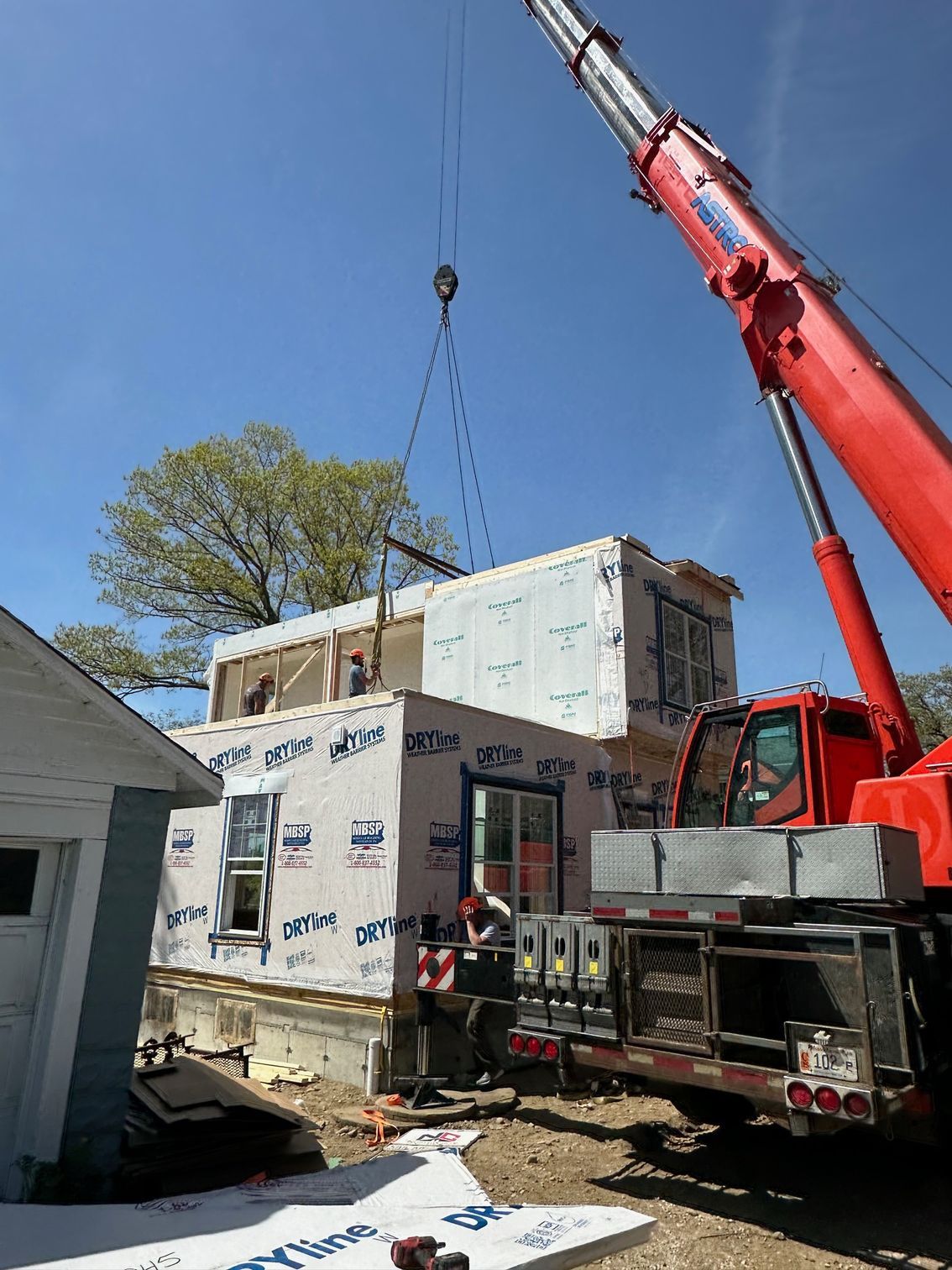 Crane lifting a prefabricated house module onto a foundation. Construction workers on site under a blue sky.