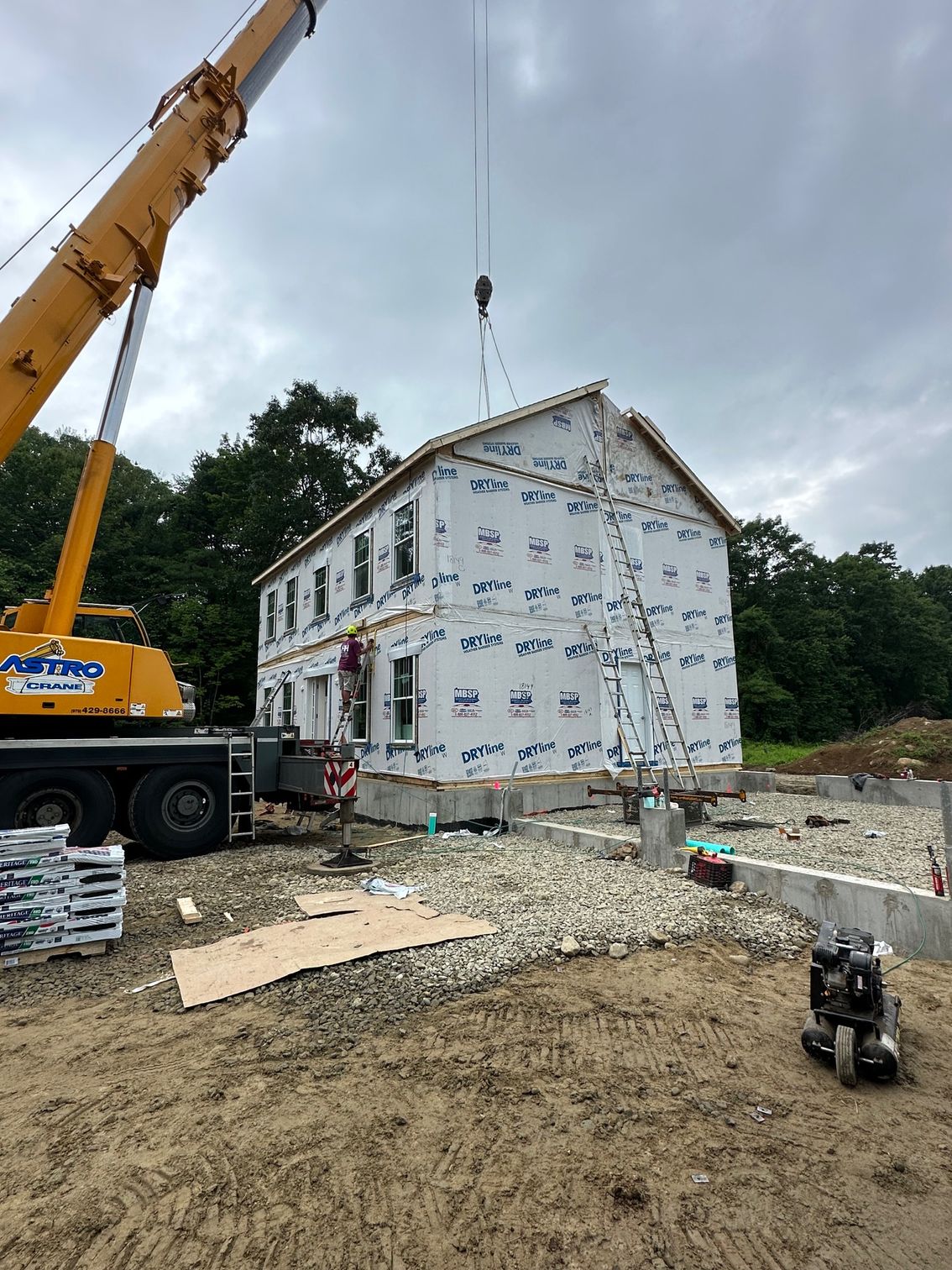 A crane lifting a modular house during construction on a gravel foundation. Blue wrap covers the walls.