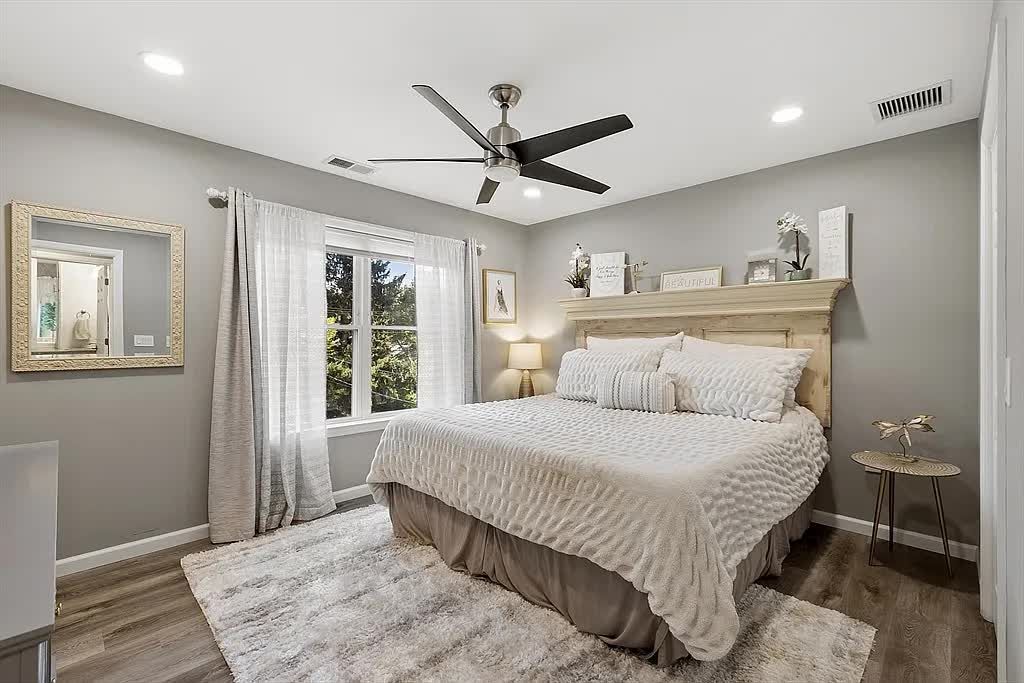 A bedroom with gray walls, wood floors, a large bed with white ruffled bedding, a wood headboard, and a ceiling fan.