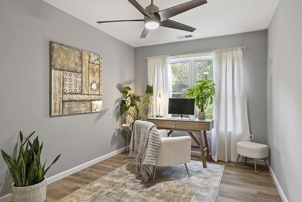 A home office with gray walls, a wooden desk with a monitor, an armchair with a throw blanket, and potted plants.
