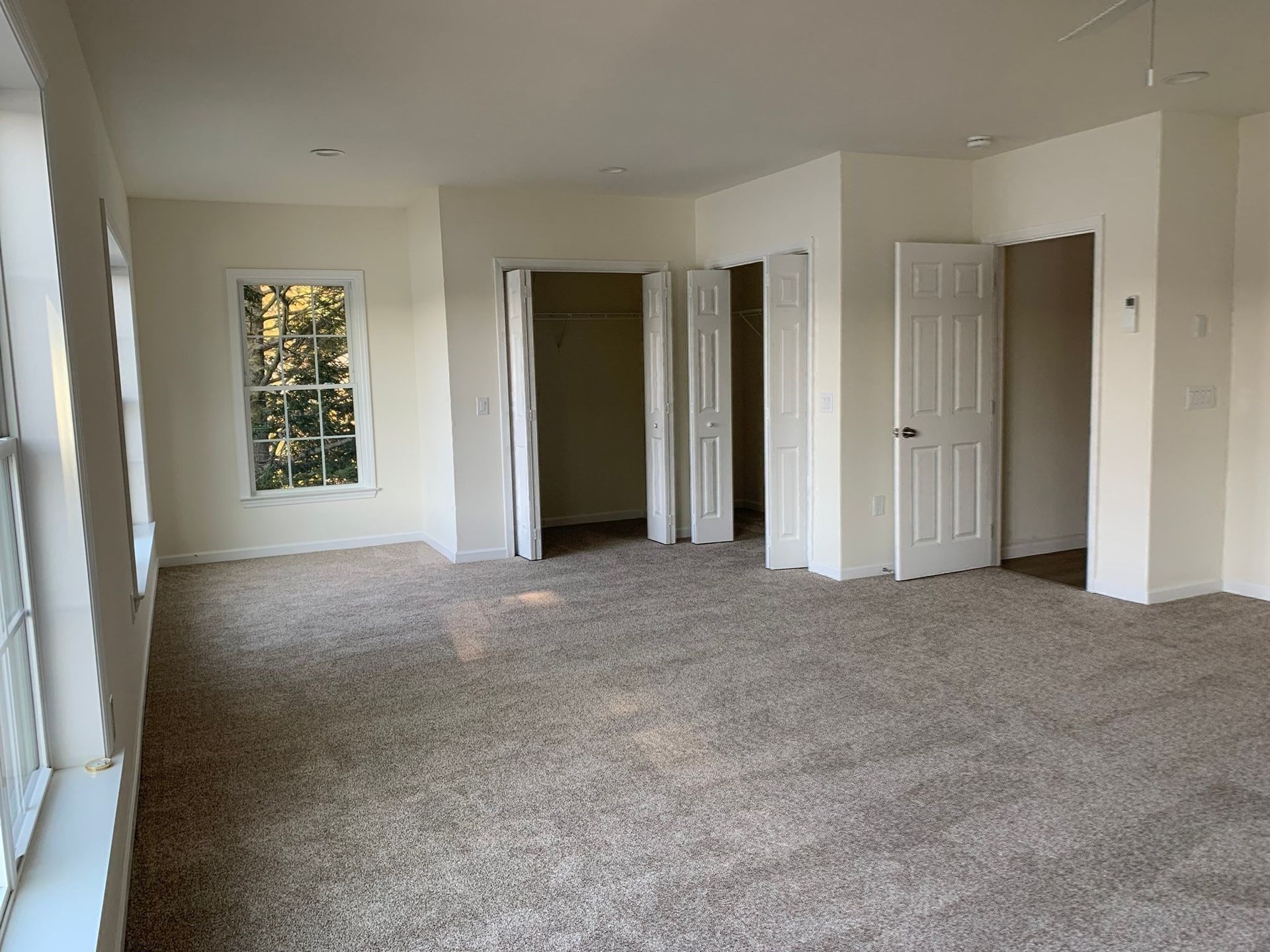 A carpeted, empty bedroom with beige walls, a window on the left, a double-door closet, and an open doorway on the right.