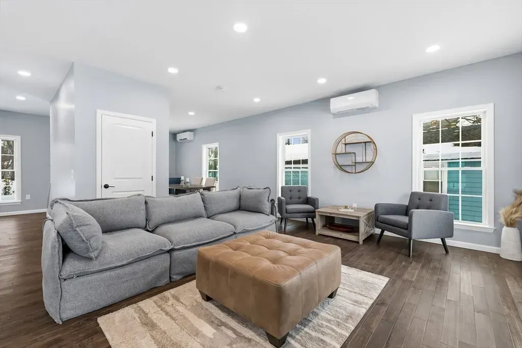 A spacious, modern living room featuring a large grey sectional sofa, a tan ottoman, two armchairs, and wood floors.