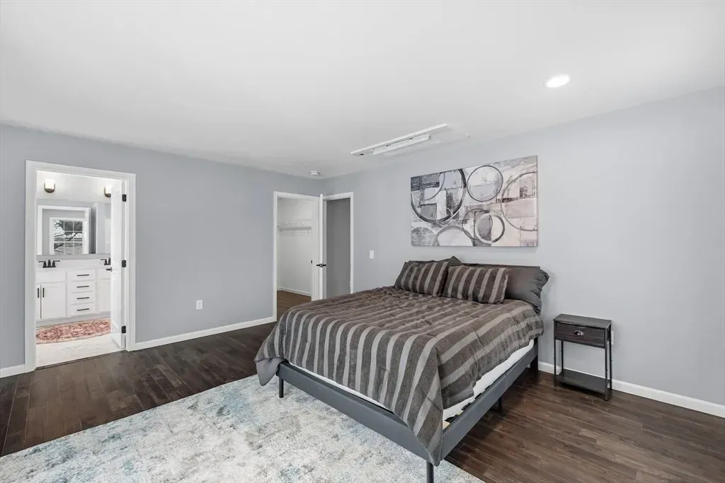 A bedroom with light gray walls, dark wood floors, a striped bed, a fluffy area rug, and an open bathroom doorway.