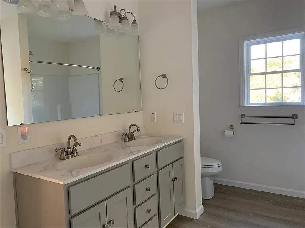 A bathroom with a dual-sink gray vanity, a large rectangular mirror, and a toilet near a window with light gray walls.