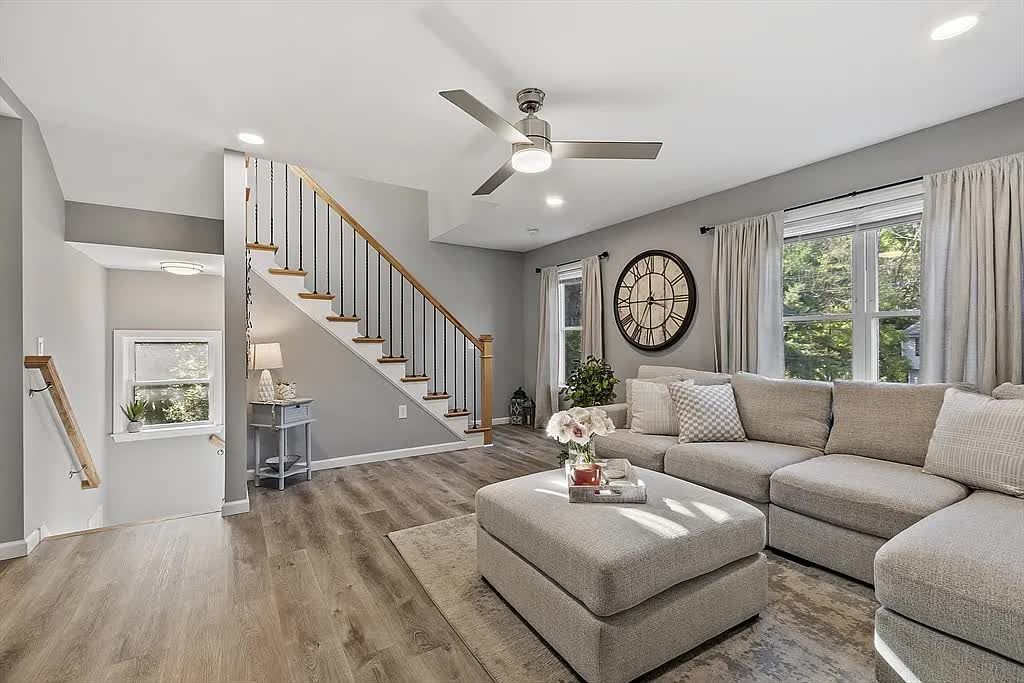 A bright living room features light grey walls, wood-look flooring, a sectional sofa, and a wooden staircase with a railing.